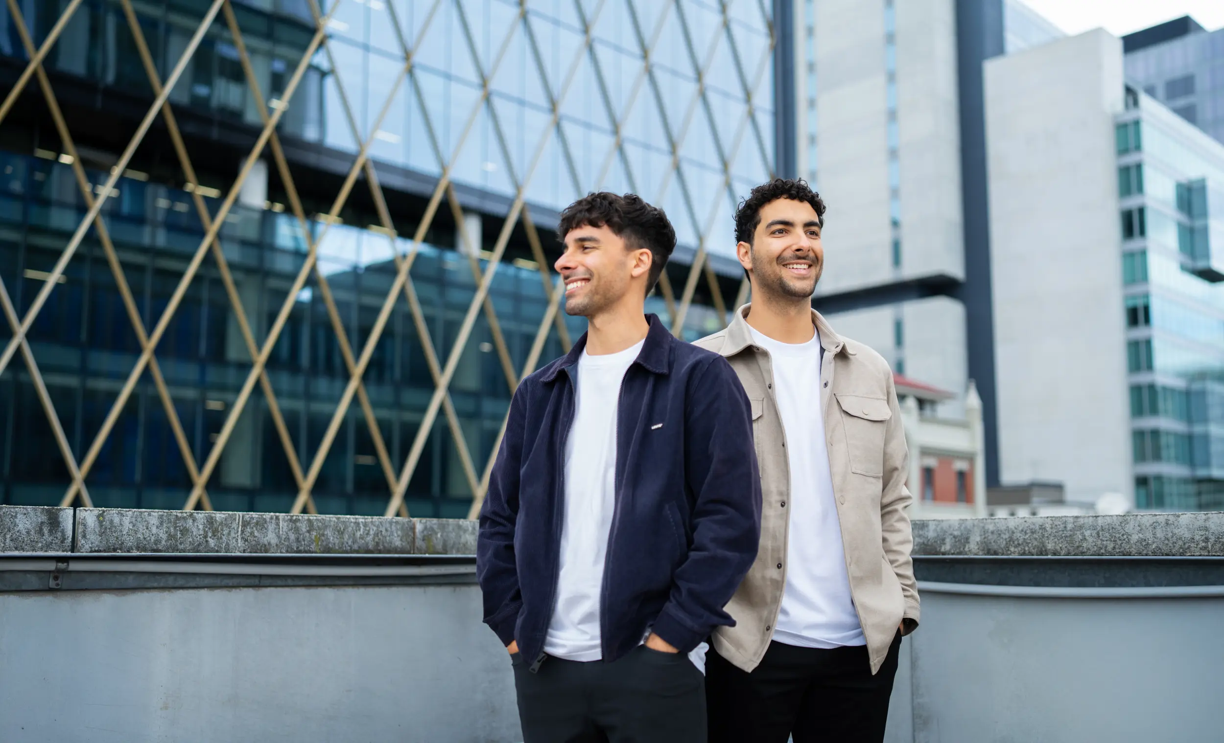 Sav and Dom smiling while looking at different directions with a skyscraper behind them.